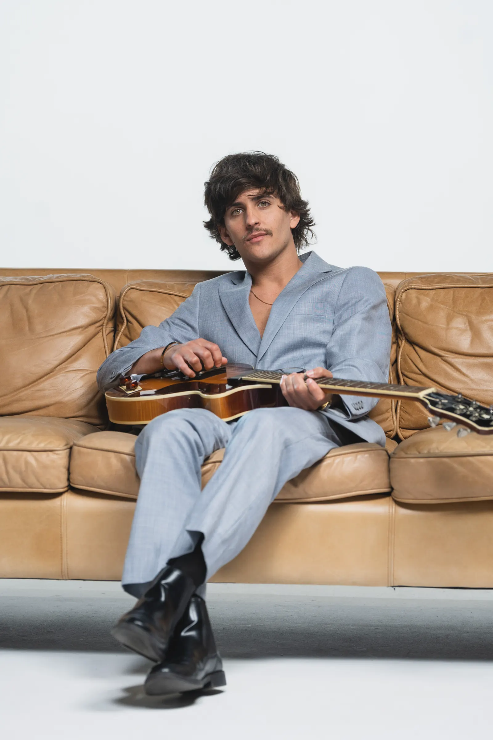 Montalvo sentado en un sofá de cuero, en traje azul cielo, sosteniendo una guitarra eléctrica